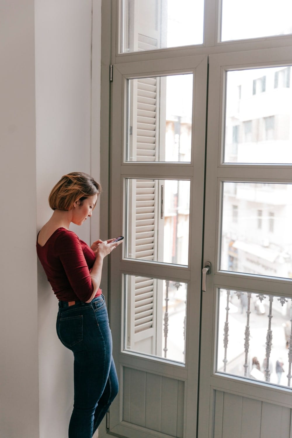 A woman stands by a large window with unique design elements, leaning against the wall as she texts on her phone.
