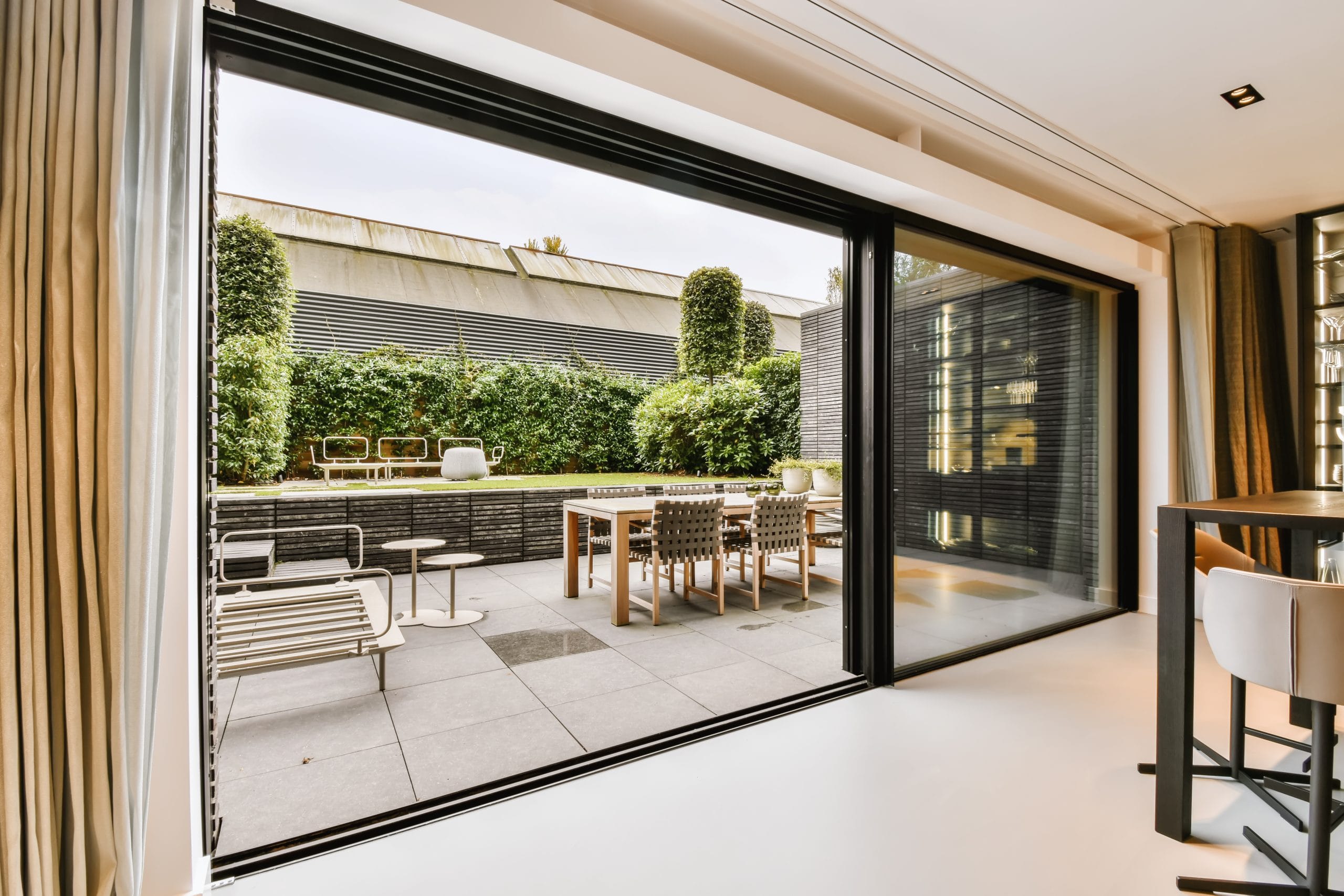 Attractive huge sliding glass door modern door installation Modern open-plan living area with glass doors leading to a stylish patio with dining table, chairs, and greenery.