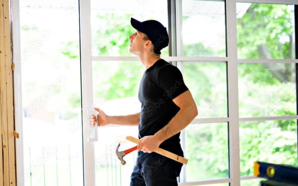 Man in black shirt and cap examines a sliding glass door while holding a hammer, with greenery visible outside.