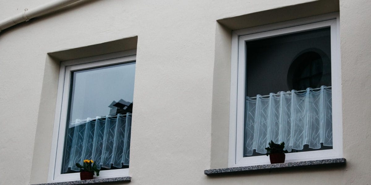Two windows with sheer curtains and small potted plants on the sills against a plain wall. Two windows with sheer curtains and small potted plants on the sills against a plain wall.
